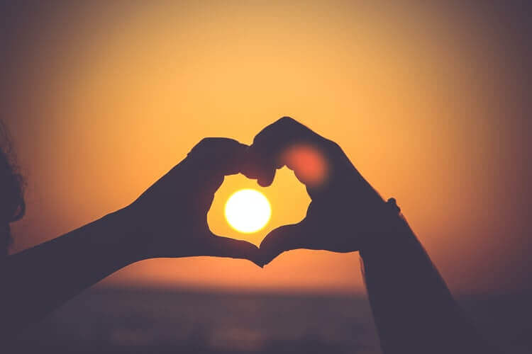 A person making a heart gesture with the setting sun in the background