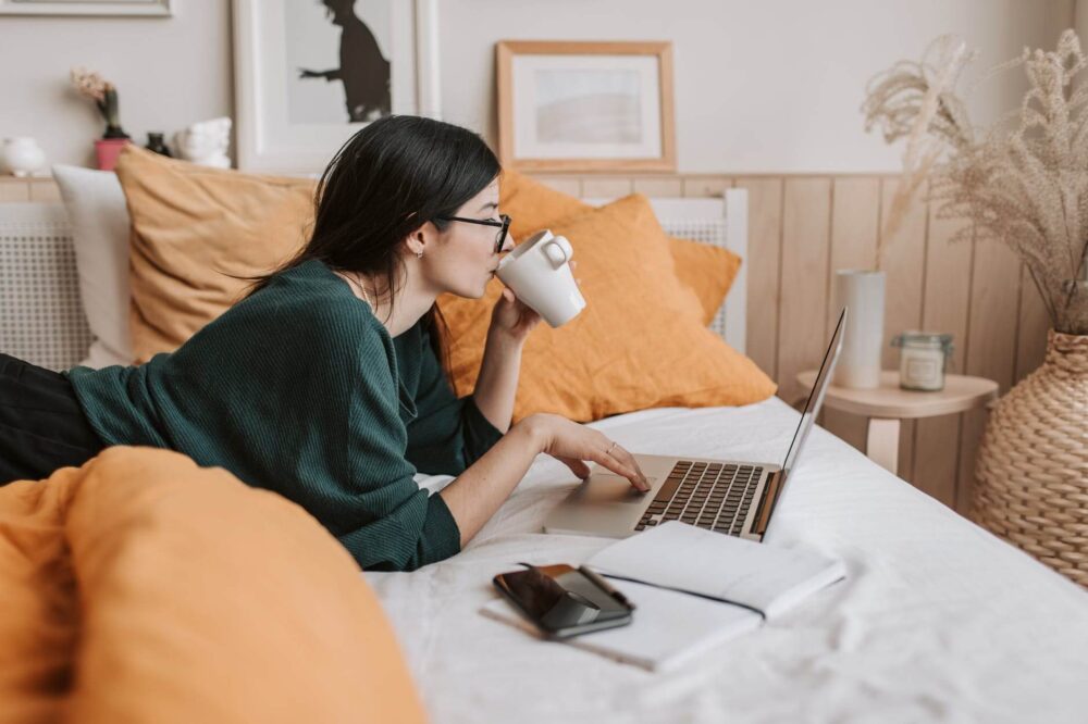 A woman using a laptop and drinking coffee while lying in bed